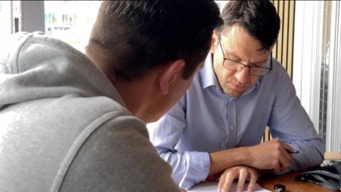 A doctor looks at paper work on his desk while a patients sits opposite him