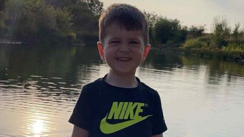 Archie York is grinning in front of a pond with the sun reflecting in the surface. He is wearing a black Nike t-shirt.