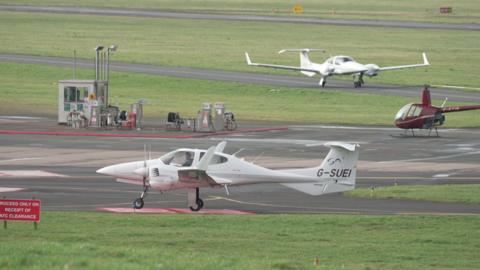 Small white propeller aircraft taxi on the tarmac of Gloucestershire airport, there is a small one seater red helicopter parked as well