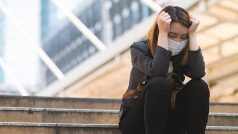 A woman wearing a mask sits on a set of steps with her head resting on her hands