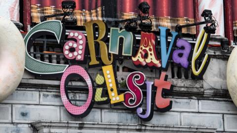 A brightly-coloured sign declaring the existence of Carnaval Aalst hangs upon a stone wall amid spotlights