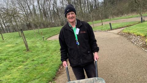 Dave Senior holding a wheelbarrow in a park area as he volunteers at Children's Hospice South West in Wraxall