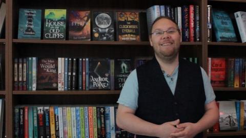 Dr Alistair Sims standing and smiling in front of books that BOTH publishing published last year