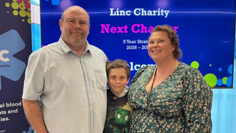 A family of three - a middle aged man and woman and their eight-year-old son, smile as they stand in front of promotional boards and screens for Linc, the blood cancer charity. The man is wearing a pale blue short-sleeved shirt and is bald, and the woman is wearing a dark green v-neck, long-sleeved dress with light orange coloured flowers on it. Their son, stood between them, is wearing a Minecraft Creeper T-Shirt. They are all smiling.
