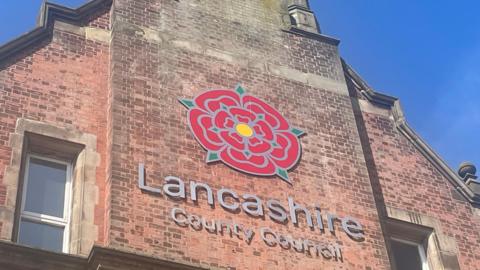 Lancashire County Council sign, with the red rose emblem, on the side of a tall, redbrick building. The photograph was taken on a sunny day with clear blue skies.