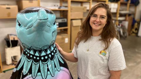 A woman wearing a grey t-shirt stands next to a teal, black and purple penguin sculpture