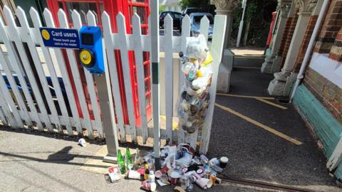Overflowing rubbish hangs from a fence at Box Hill & Westhumble railway station on 5 July 2026. Plastic bottles, coffee cups, cans and packaging have spilled onto the ground beside a ticket validator and station entrance.