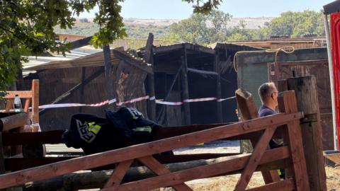 Fire-damaged farm building cordoned off with safety tape, viewed behind a wooden gate. A firefighter's jacket rests on the gate, with a person standing nearby and scorched structures visible in the background.