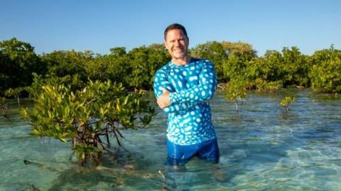 Steve Backshall wearing a blue waterproof short and shorts standing thigh-high in crystal clear water