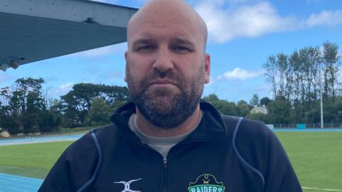 Jordan Reynolds has no hair and has a short dark beard. He is looking into the camera.