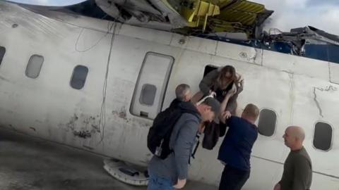 A woman is helped through a plane door by two men on the tarmac. The plane is upside down