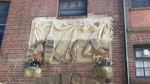 A sandstone frieze is impressed in a brisk wall. It depicts two people in profile facing each. One playing a stylized image of an instrument that looks like a lute and the other playing what appears to be a saxophone. Below the frieze are two sandstone bowls with coloured flowers.