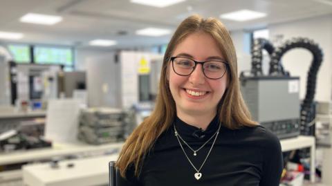 Amy Gilmore is standing in a mechanics lab and smiling at the camera. She has long brown hair and is wearing black glasses.