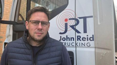 The photo shows Oliver Reid looking at the camera. He is wearing a navy gilet and jumper. He has short brown hair and is wearing glasses. He is standing in front of a white HGV lorry, which has the logo 'John Reid Trucking' on it. 