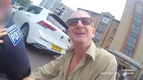 Body-worn camera image of a smiling Jeff Stewart squatting behind a policeman in a car park. He is smiling and is wearing sunglasses and a pale shirt with several buttons open at the chest.