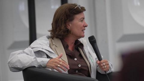 Eluned Morgan wearing a white coat, beige jacket and red top, holding a microphone to her mouth.