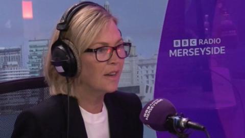 Paula Basnett, wearing glasses and a blue suit jacket and white top, speaks into a purple microphone in BBC Radio Merseyside's studio