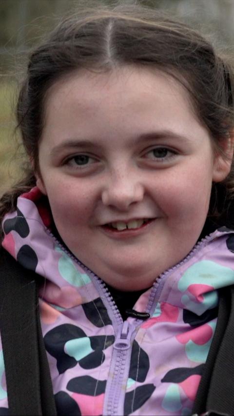 A smiling girl with dark brown hair looks at the camera. She is wearing a purple coat with a black, pink and turquoise pattern on it. 
