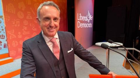 Alex Cole-Hamilton smiling straight at the camera. He is wearing a dark suit over a light shirt with a salmon pink tie. He is standing at a lectern with two black microphones coming out the top. He is standing on a stage in front of an orange background. The Liberal Democrats' party logo is over his left shoulder in a white font.