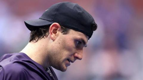 A white man wearing a black cap backwards. He has a plain expression on his face and facing down. We are looking at his side profile.