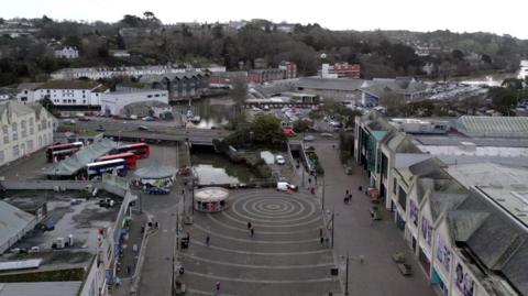 Aerial view of a Truro city centre with circular patterns on the ground, surrounded by buildings, buses and a river with bridges in the background. White skies.