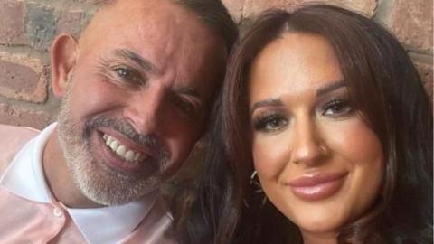 Billy Morley and his wife Ashley have their heads pushed together and are smiling brightly at the camera. Mr Morley has short, dark hair and a grey beard. 