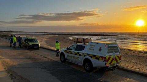 Two rescue vehicles parked along the shoreline with a four men dressed in high visibility clothing with one looking out to see through binoculars. It is late evening and the sun is setting over the sea.