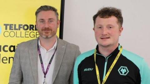 Two men stand to the right of a yellow screen which has the Wolves Foundation and Telford College logos on it. The man on the left is wearing a checked blazer and white shirt and the other a Wolves black and turquoise tracksuit. Both have name badge lanyards on and are smiling at the camera.