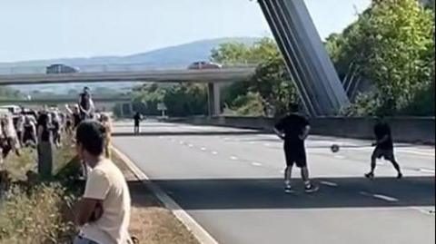 A blurred screenshot shows a group of people sat on the central reservation on a closed motorway. On the right, two people can be seen kicking a ball between them on an empty motorway, beneath a bridge. Another person can be seen walking in the background. 