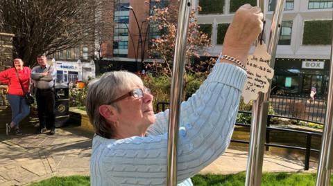 A woman attaches a wooden sign that reads "Jim Allen, taken too soon, always in my heart xx" to a metal structure.