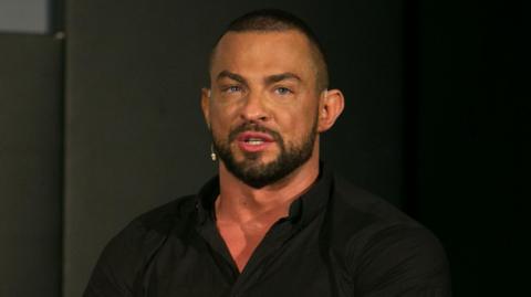 Robin Windsor wearing a collared black shirt. He has an earpiece and a beard. He is looking directly at the camera.