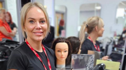 A blonde woman wearing a lanyard smiles at the camera. She is standing on front of a mirror which has a training mannequin head in front of it.