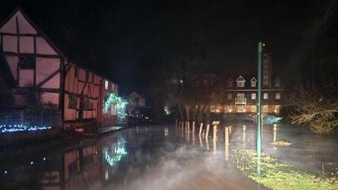 A flooded road seen in the dark with wooden posts clearly seen which would normally mark the road from the river bank but surrounded by water. There are houses on the side of the road with Christmas lights on the outside.