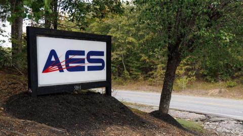 A sign reading AES is posted on the side of a rural roadway surrounded by trees and other greenery.