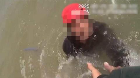 A blurred out face in water, he is wearing a black wetsuit and a red cap. There is a hand reaching out to get him out of the water.