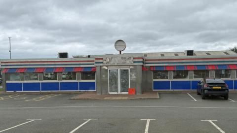 The former OK Diner building has the windows scrubbed and is in the process of shutting. It is a long single-storey concrete building with large windows and red and blue branding, in the style of a traditional 1950s American diner. A single black car is parked outside, to the right of the central entrance.
