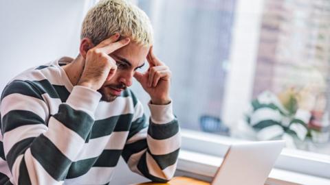 A young man with short bleached blonde hair and a beard, wearing a striped long-sleeved T-shirt, sits at a desk, looking toward an open laptop. He looks frustrated and is resting his fingers against the sides of the forehead. A large window and houseplants are visible behind him. 