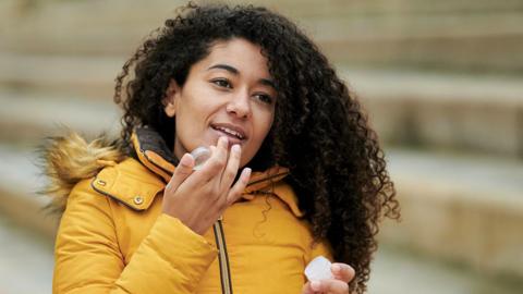 A woman outside in winter is wearing a bight yellow coat and applying lip balm to her lips. 