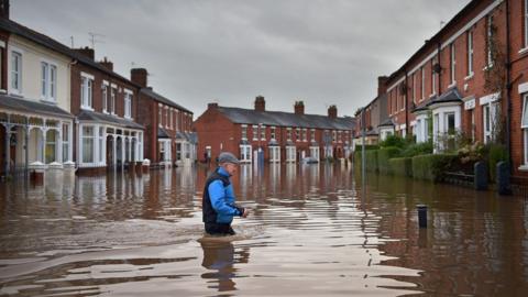 A Carlisle resident makes his way through floodwater. Red brick terraced houses on both sides of the road are submerged.