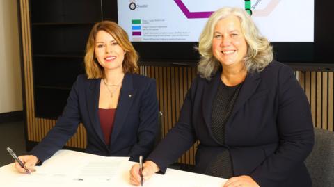 Kim McGuinness and Heidi Alexander sit side by side at a desk while signing the document. They are both looking at the camera and smiling. McGuinness, on the left, has mid-length blonde hair and is wearing a blue blazer. Alexander has mid-length light blonde/grey hair and is wearing a blue blazer.
