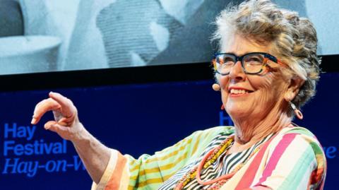 A woman with short grey hair wearing navy and orange glasses and a multicoloured stripey dress. She has a microphone on her cheek and is talking.