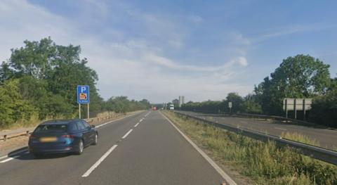 The A1. The road, which stretches into the distance, is almost empty. Each carriageway has two lanes. There is a blue estate car in the foreground on the left of the picture, heading away from the camera. In the far distance, heading towards the camera, there is white lorry. It is a clear day and there are leaves on the trees beside the road.