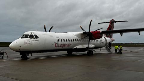 A Loganair plane on the tarmac at Jersey Airport on a cloudy day. The aircraft is white and red. It has black twin propellers. Two people in high-vis jackets are at the back of the plane.