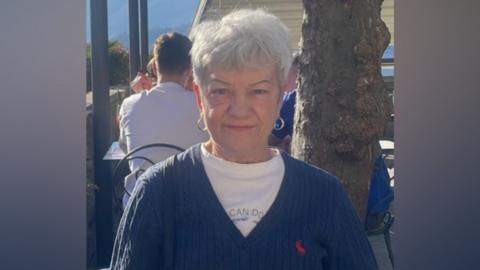 A woman with short grey hair sits at an outdoor café, wearing a navy cardigan over a light-coloured top. A tree trunk is beside the table, with other customers seated in the background.