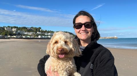 Annabel Hill wears dark sunglasses and a black fleece jacket and stands on a sandy beach holding a small, curly‑haired dog. The dog has light-coloured fur and its tongue is visible. Behind them is a calm sea, a clear blue sky with scattered clouds, and white buildings on a hillside. A pier extends into the water in the distance.