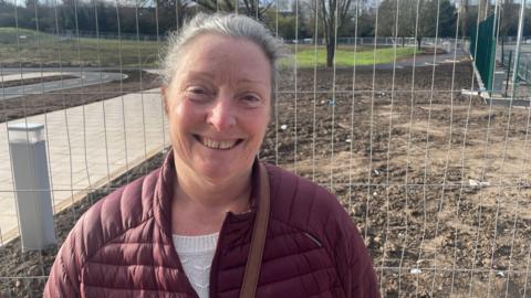 A smiling woman standing outside in front of metal construction fencing, with bare earth and a park behind her. She is wearing a burgundy quilted jacket over a white top and has her hair tied back. The sun is shining on her face.