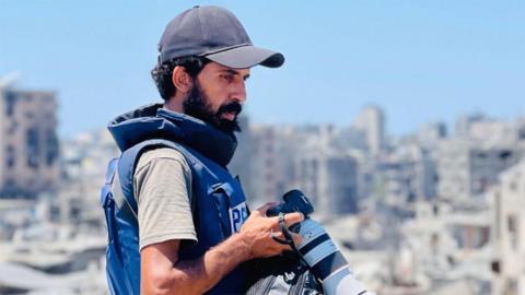 A man with a beard, wearing a baseball cap, holds a professional-standard camera. He is in profile and wearing a flak jacket with the word "Press" on it, and he appears to be standing on the upper level of a building on a sunny day. In the background is the blurred image of what appears to be a heavily bomb-hit city.