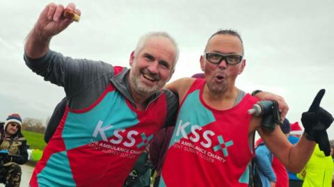 Two men wearing red and light blue vests with the letters KSS on the front. They are smiling at the camera. Behind them is a grey sky