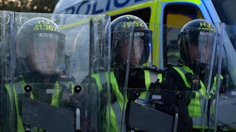 Three police officers in riot gear holding riot shields in front of them.
