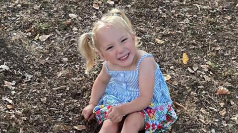 Bella smiles and looks up at the camera while she sits on the floor. She has blonde hair which has been tied up in pigtails. She is wearing a blue and white striped summer dress. 